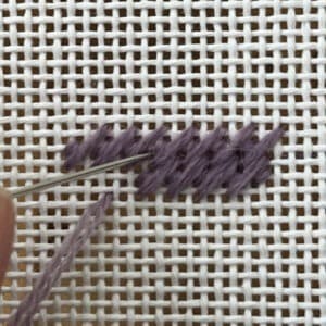 Close-up of a purple embroidery thread stitching through white fabric in a cross-stitch pattern, with a needle passing through the threads.