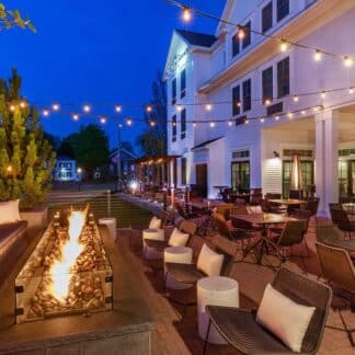Outdoor hotel patio at dusk with a long fire pit, cushioned lounge seating, and string lights above white buildings.
