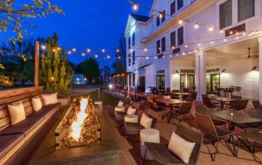 Outdoor hotel patio at dusk with a long fire pit, cushioned lounge seating, and string lights above white buildings.