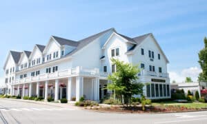 White multi-story hotel building with a wraparound porch, gabled roofs, and well-kept landscaping under a blue sky; a small tree and flower beds line the front.