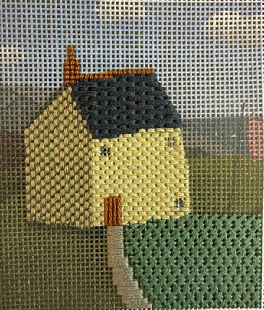 Needlepoint  of a small yellow house with a brown roof on a grassy hill.